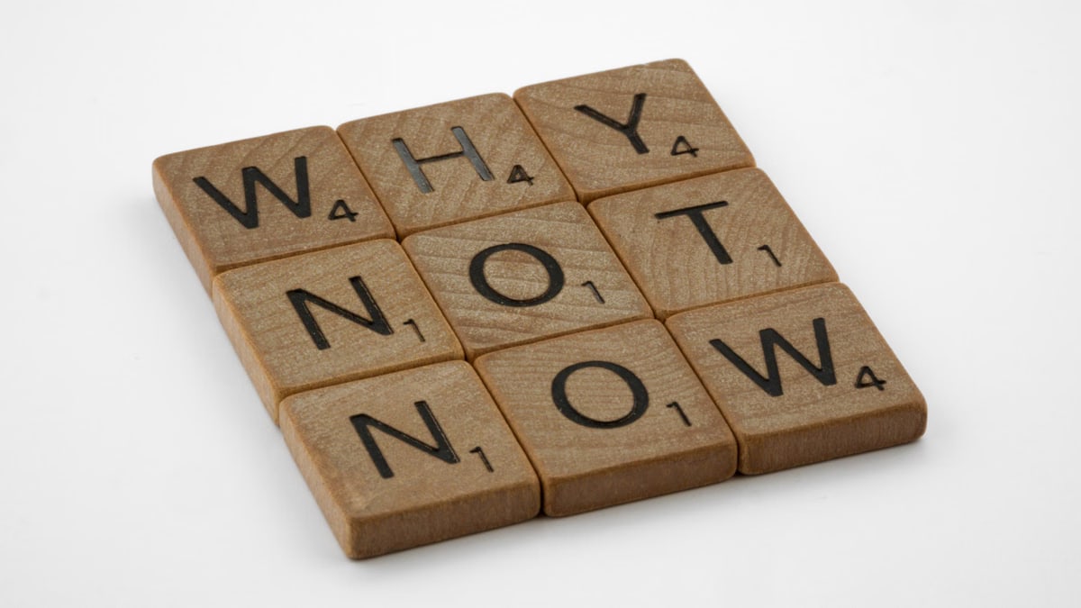 Wooden letter tiles form the motivating phrase 'Why Not Now' on a white background, encouraging action and decision-making.